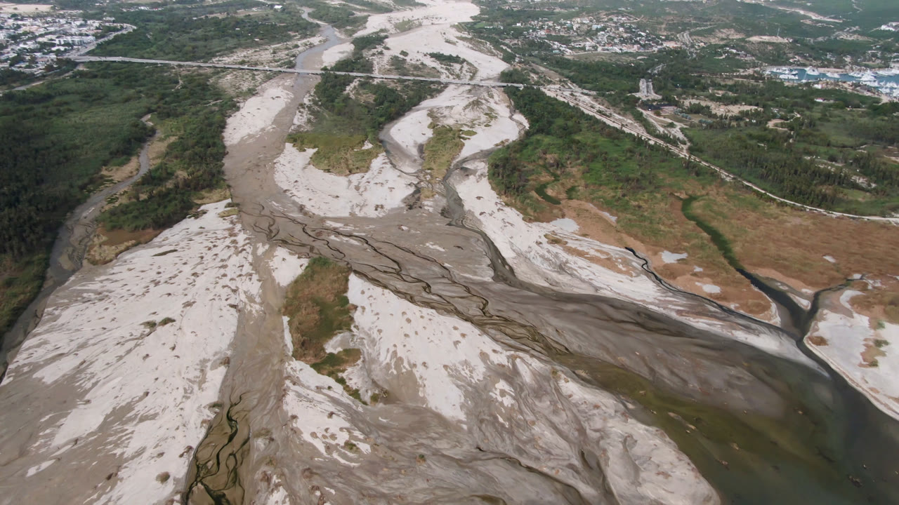 Aerial view over a braided river valley in San Jose Del Cabo, Baja California Sur, with pale sandbars, green scrub, distant buildings, and no visible people.