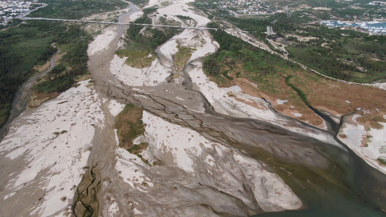 High aerial frame of San Jose Del Cabo river wash showing gray erosion channels across white sand, green vegetation along the banks, roads, and no visible people.