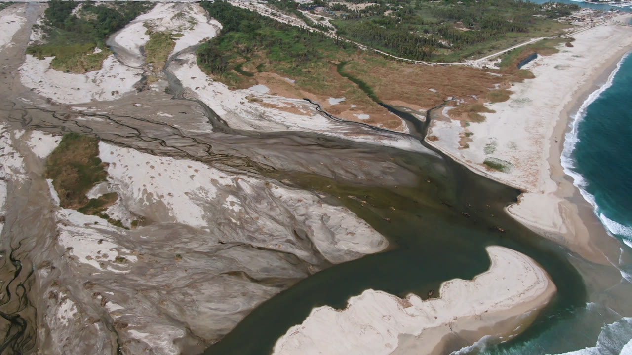 Aerial frame from San Jose del Cabo shows a dark river channel dividing pale sand banks before reaching blue ocean surf at the right, with no visible people.