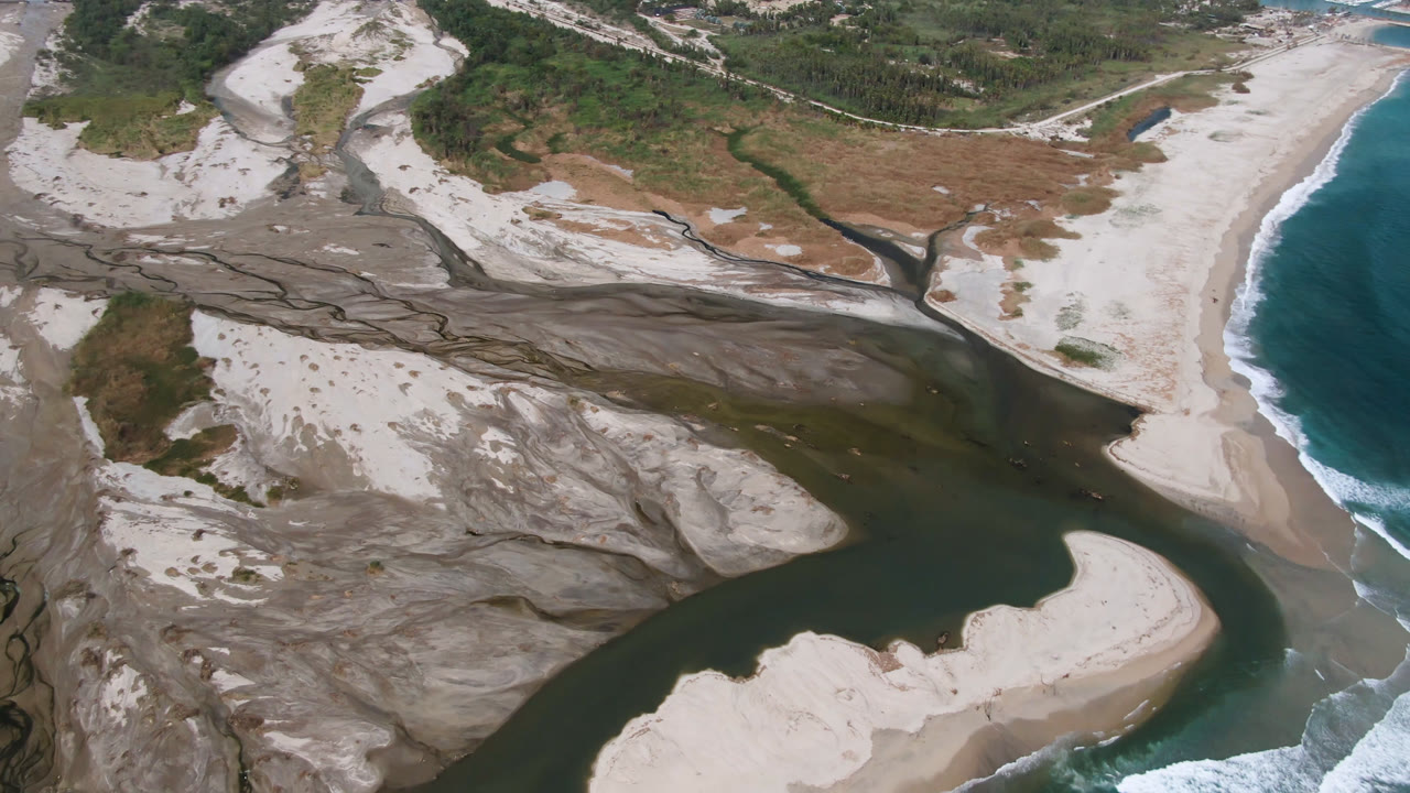 Aerial frame from San Jose del Cabo shows braided gray erosion channels feeding a darker river beside pale sand bars, with ocean surf along the right edge and no visible people.