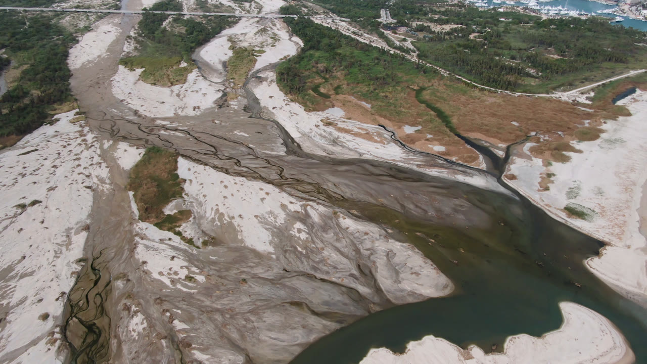 Aerial frame over San Jose del Cabo showing a dark river bend below gray erosion channels and pale sand, with scrubland beyond and no visible people.