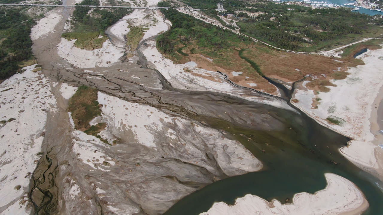Overhead view in San Jose del Cabo with braided dry washes feeding toward a dark river channel, bordered by pale sandbars and green scrub, with no visible people.
