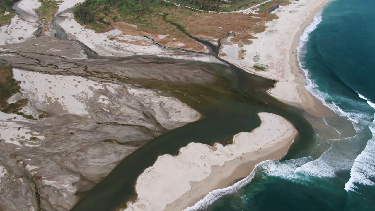 Overhead view in San Jose del Cabo, Baja California Sur, showing a dark river channel bending around pale sand banks toward teal ocean waves, with no visible people.