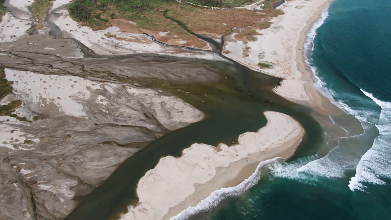 Aerial frame over San Jose del Cabo with the river inlet centered, a bright sand bar in the lower frame, white surf curling on the right, and no visible people.