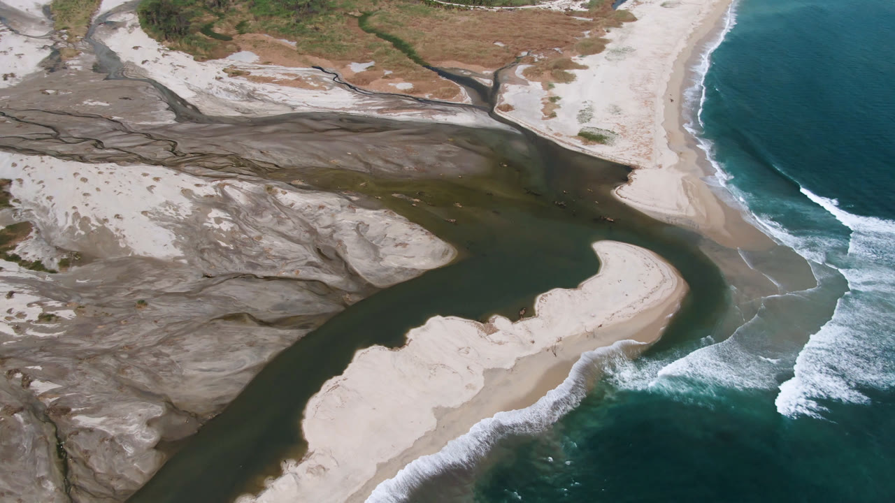 Drone view of river water cutting through sand banks beside breaking surf.