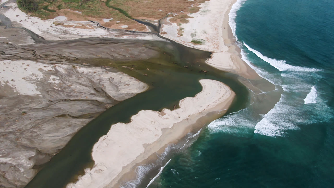 Overhead view in San Jose del Cabo of a dark river channel curving around pale sandbanks toward blue-green surf, with no visible people.