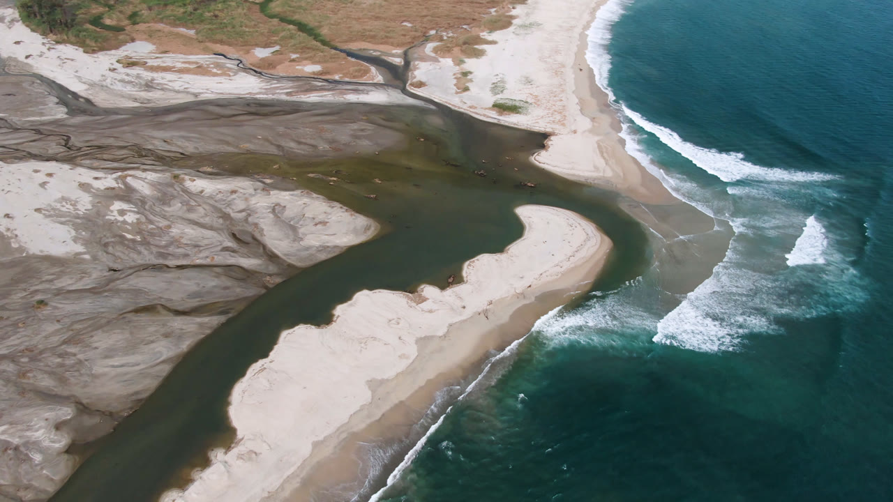 High aerial frame in San Jose del Cabo with the river running diagonally past a pale sandbar as white waves break along the beach, with no visible people.