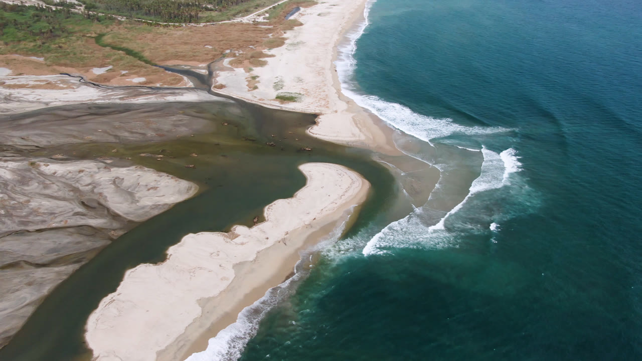 Aerial frame from San Jose del Cabo showing a dark green river channel curving through pale sandbars toward teal ocean waves, with no visible people.