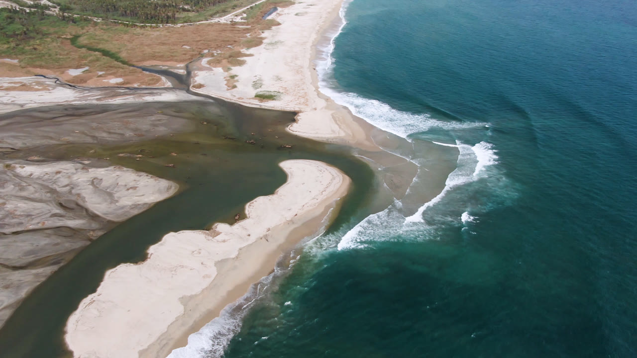 Aerial frame from San Jose del Cabo with a curved sand spit between the river channel and ocean, while white waves break along the right shoreline and no visible people appear.