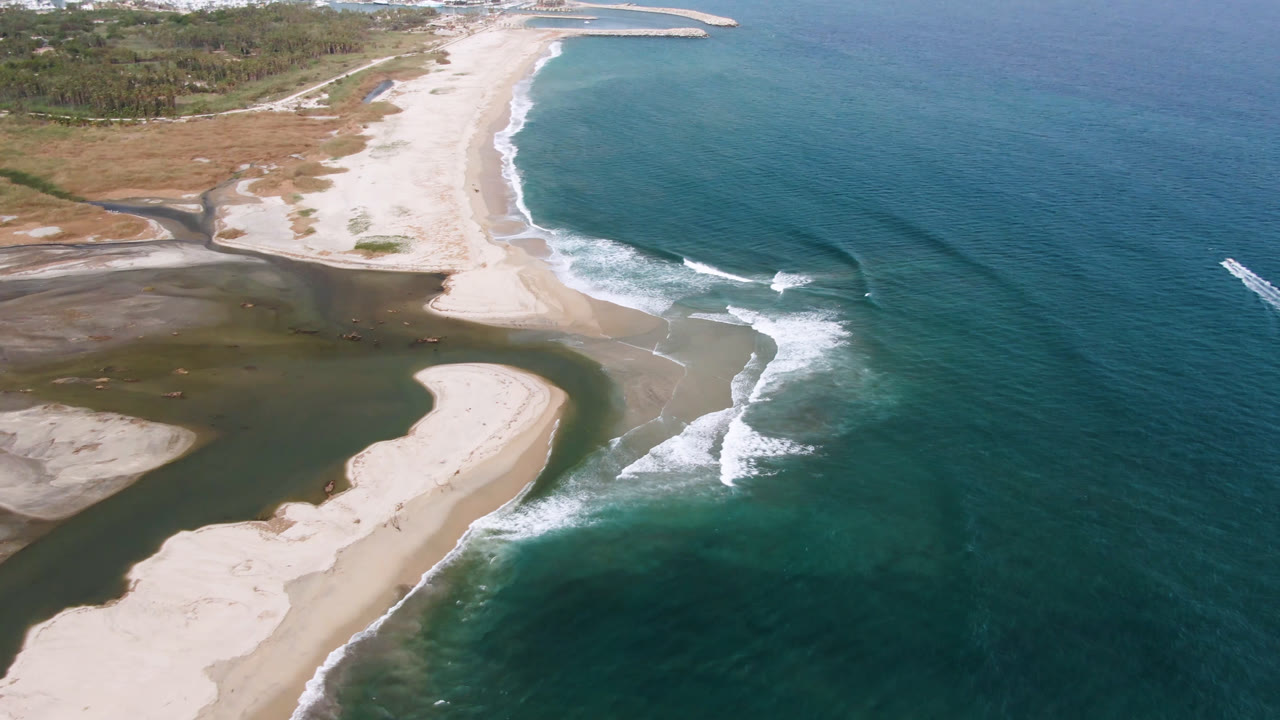 Aerial frame from San Jose del Cabo showing pale sandbars around a dark inlet, turquoise ocean, white surf, coastal vegetation, and no visible people.