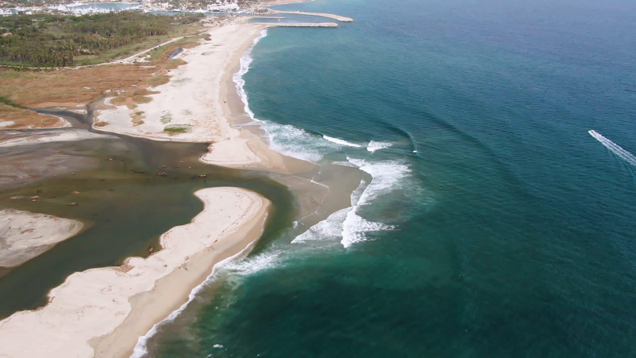 Aerial view over San Jose del Cabo with the curved sandbar more centered, surf breaking along the beach, a distant breakwater, and no visible people.