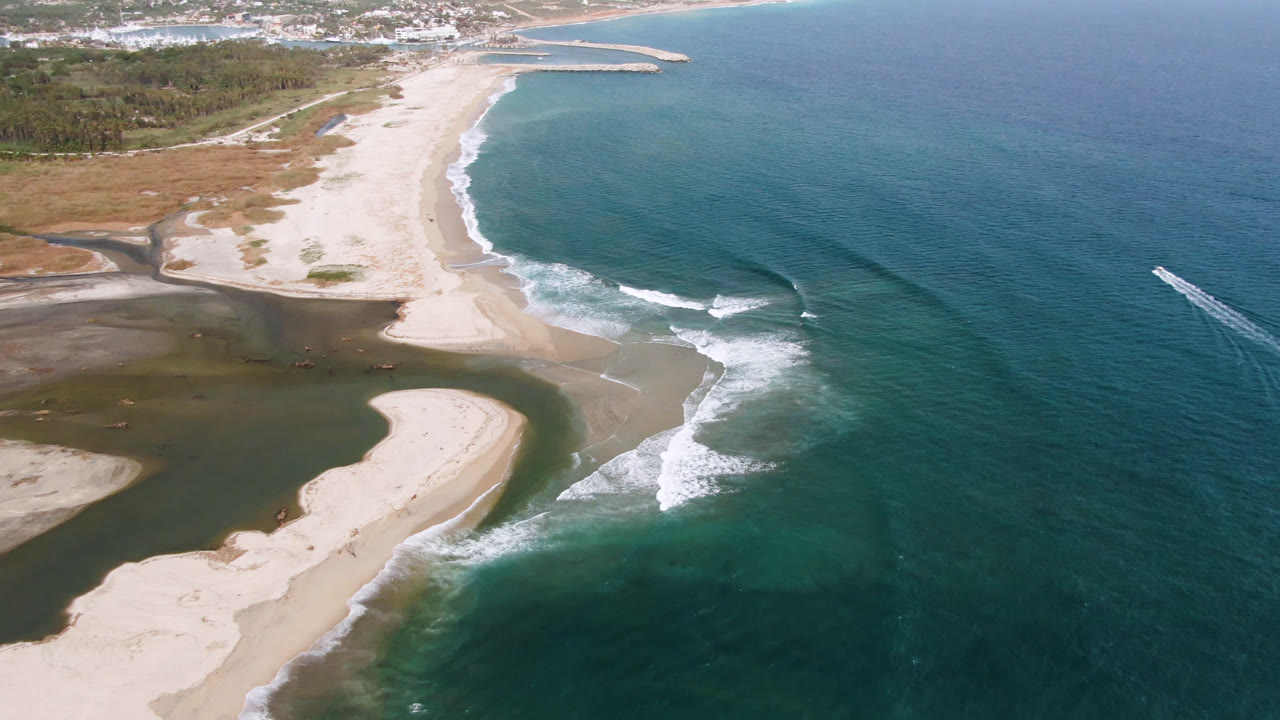 High aerial of beach, inlet, breaking waves, and a distant harbor breakwater.