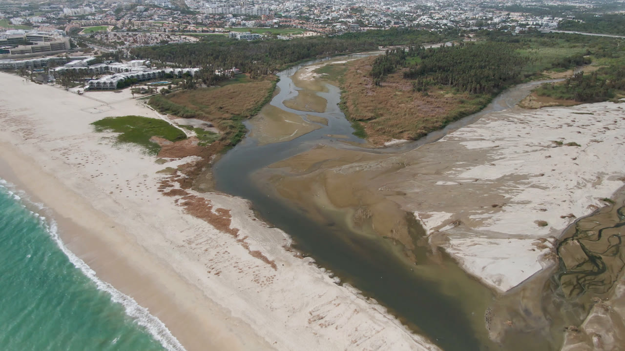 Aerial frame over San Jose del Cabo showing a dark river channel reaching pale beach sand beside turquoise surf, with marsh vegetation and city development in the distance, no visible people.