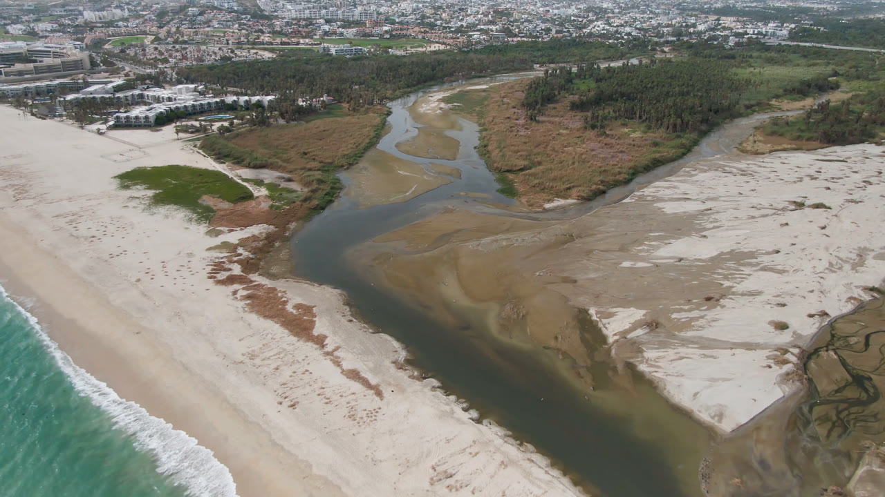 Aerial view in San Jose del Cabo with the river delta centered between pale sand flats, green marsh patches, beach surf at lower right, and buildings set back near the horizon, no visible people.