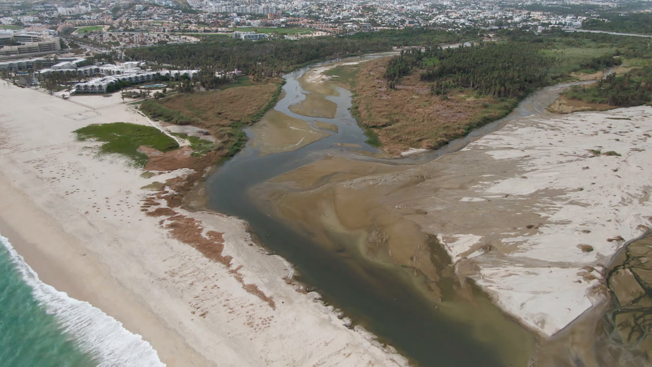 Aerial coastal scene with a river channel, sand banks, surf, and city beyond.