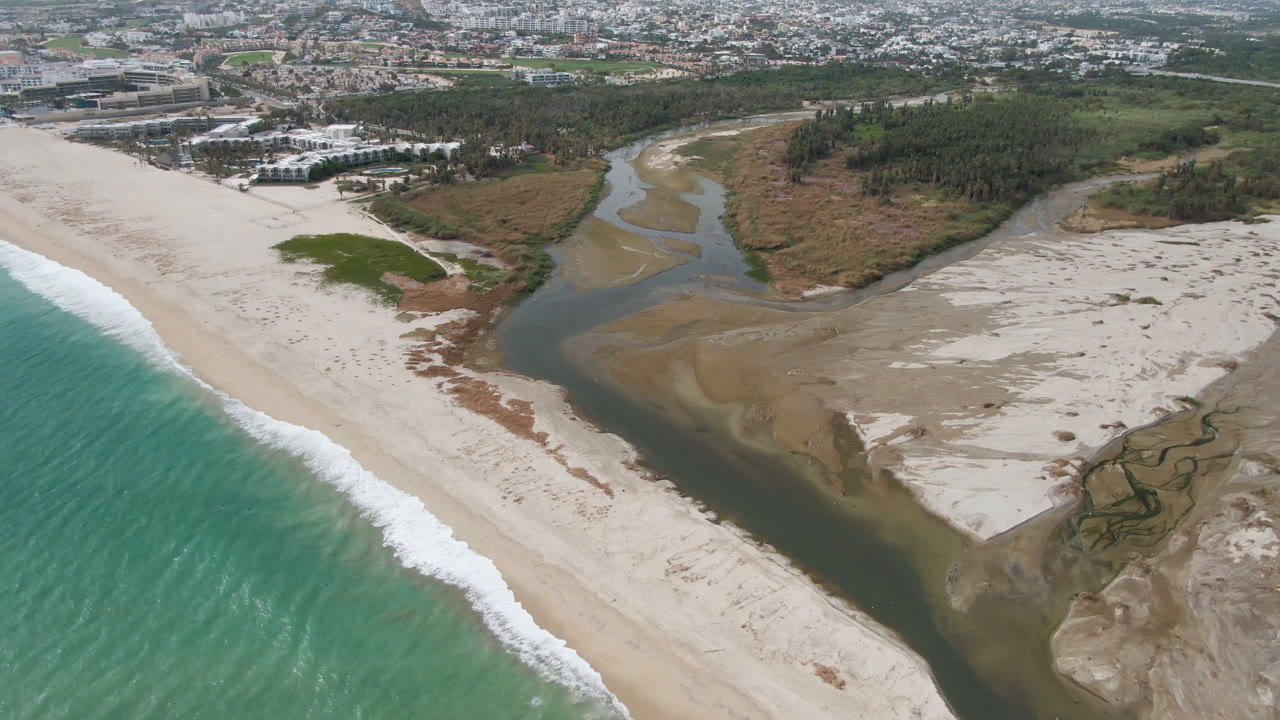 Opening aerial frame over San Jose del Cabo shows turquoise surf beside a pale beach, a dark river channel crossing sandbanks, marsh textures, trees, and distant buildings with no visible people.