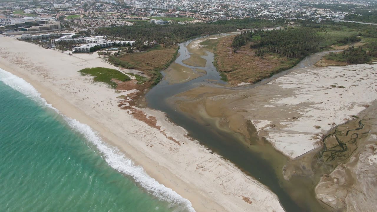 Aerial coastal frame with ocean waves, broad beach, river inlet, sandbars, wetland textures, and town beyond.