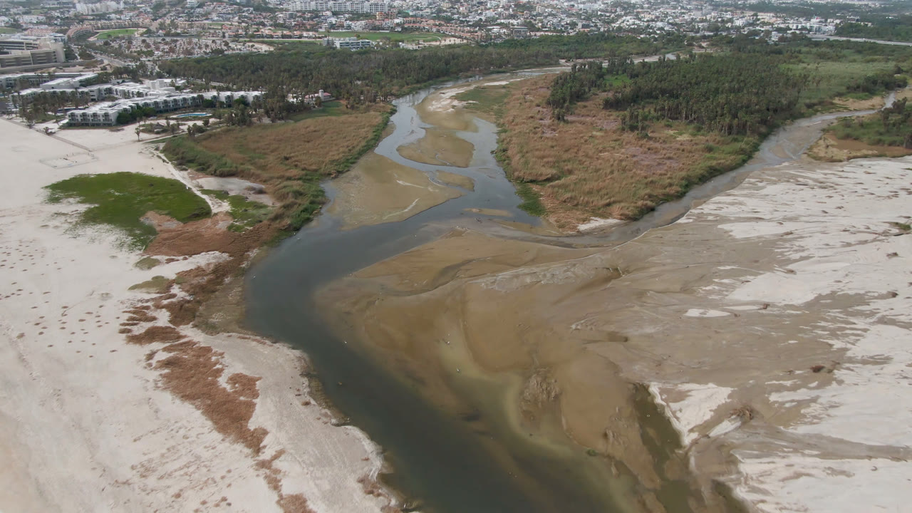 Aerial view in San Jose del Cabo of a dark river channel curving through pale sand flats, with green vegetation patches, a tree line, distant buildings, and no visible people.
