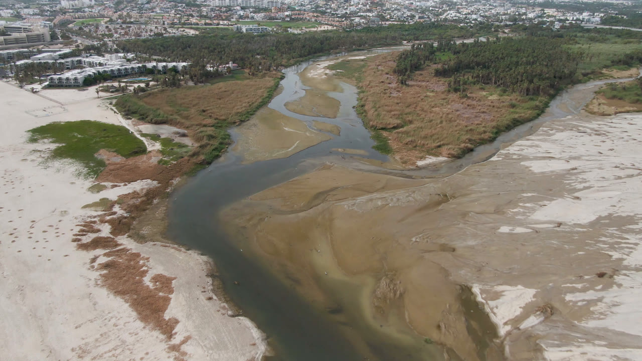 Aerial frame over San Jose del Cabo with the river bend centered between tan sandbars, brushy vegetation, distant city blocks, roads, and no visible people.