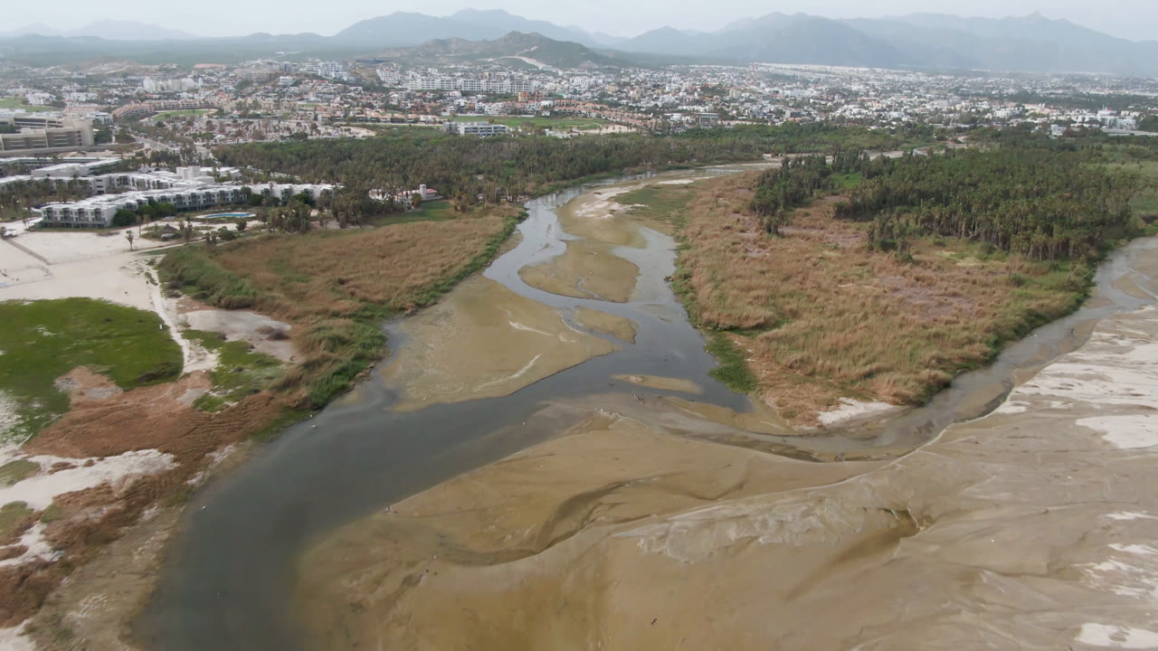 Aerial view in San Jose del Cabo showing a winding river across sandy flats, dry grass, green vegetation, distant city buildings, hazy mountains, and no visible people.