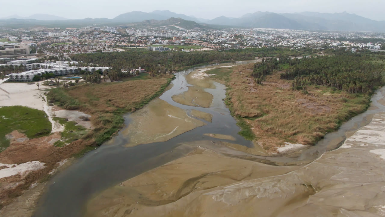 Drone view in San Jose del Cabo with the river bending through pale sandbars, dry grassland, a tree belt, low city buildings, hazy mountains, and no visible people.