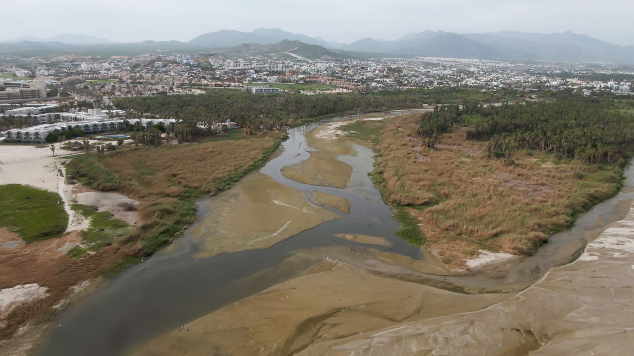 Wide aerial landscape with a river channel, dry field, trees, city, and mountains.