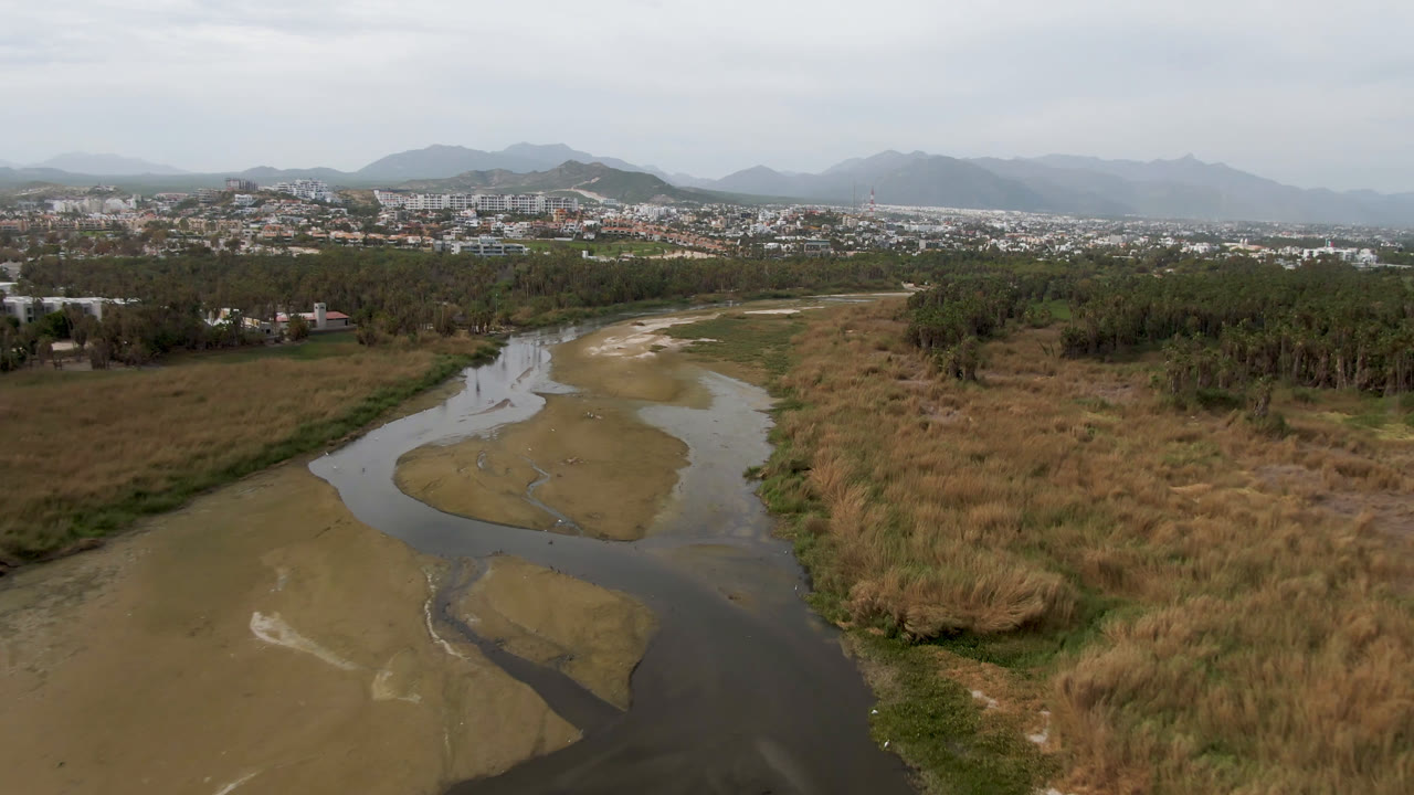 Drone view of a river bending through dry grassland toward a distant city and mountains.