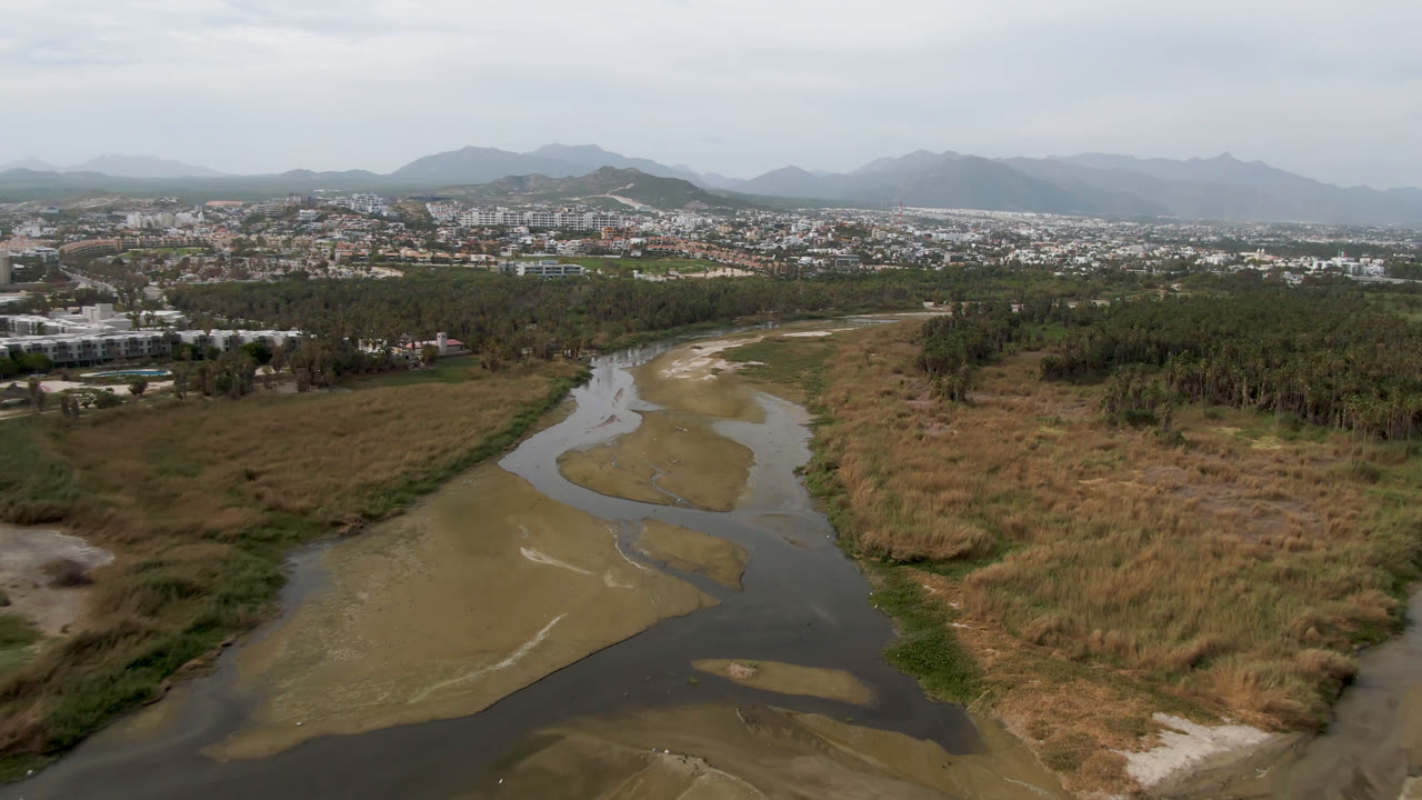 Aerial frame from San Jose del Cabo showing a muddy river bending through marshy vegetation, with a tree belt, city edge, hazy mountains, pale sky, and no visible people.