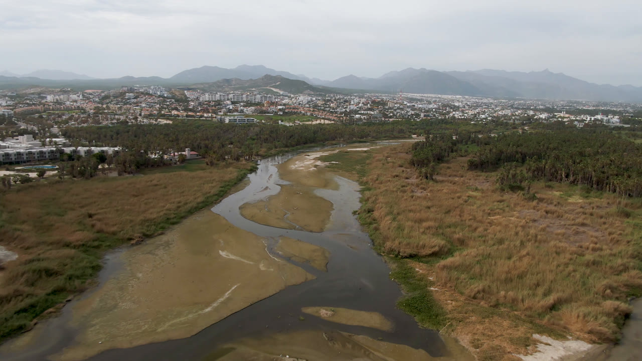 Aerial frame from San Jose del Cabo with the river channel centered between tan grasses and darker green vegetation, backed by low city development and distant mountains, with no visible people.