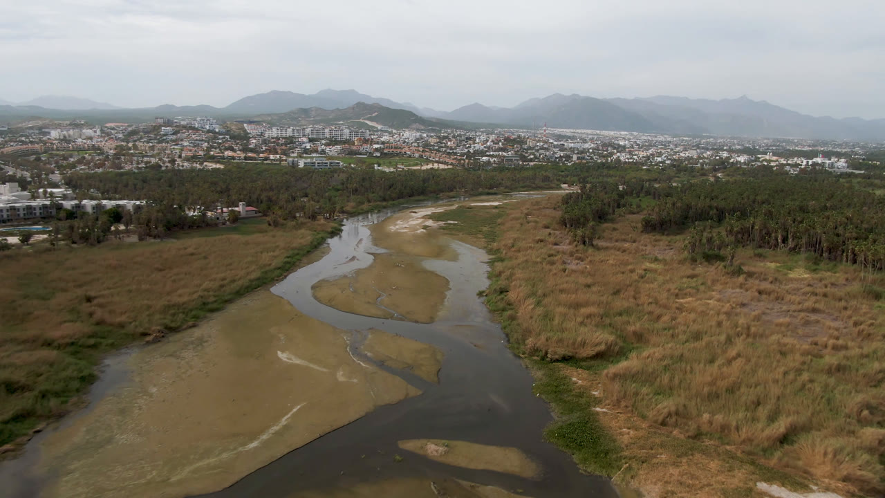 Wide aerial landscape of a river bend, marsh vegetation, city edge, and mountain backdrop.