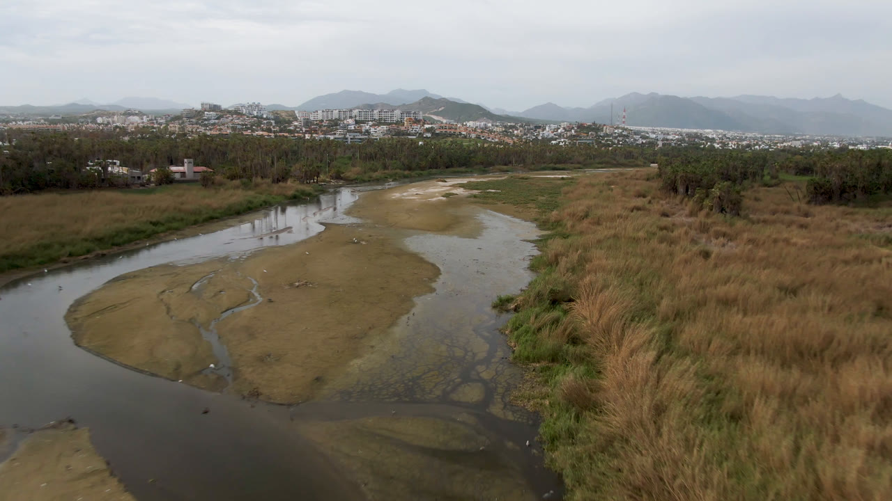 Aerial frame from San Jose del Cabo showing a shallow river curving through marsh grass and exposed sandbars, with distant buildings and mountains under a pale sky, no visible people.
