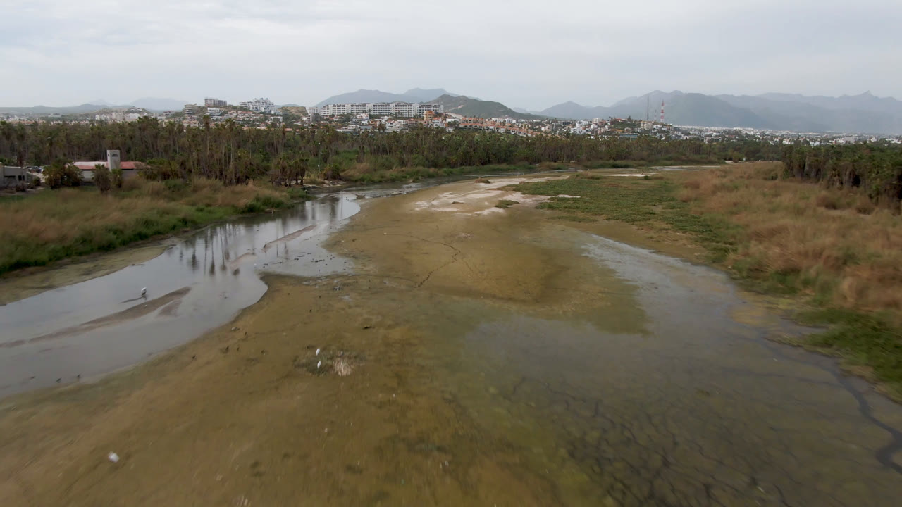 San Jose del Cabo aerial frame with the river bending left through exposed sandbars, rough grass, a line of trees, low buildings in the distance, hazy hills, and no visible people.