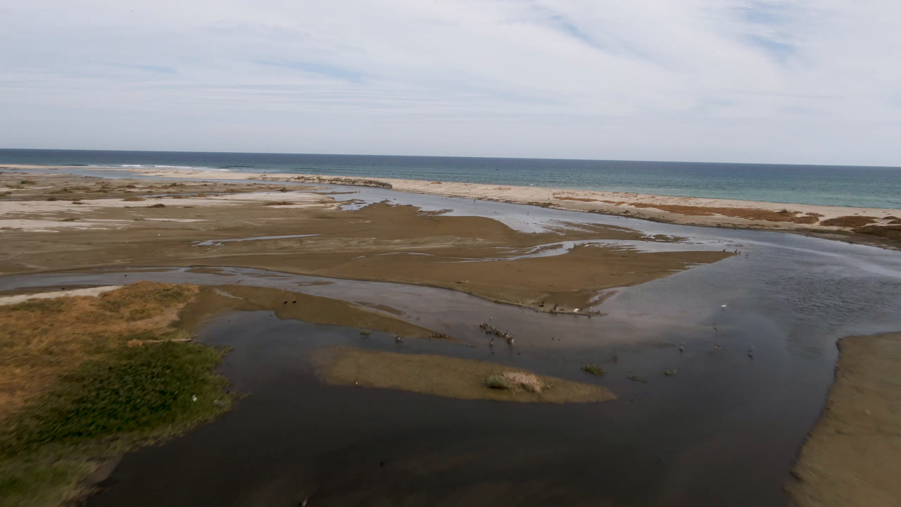 Wide aerial frame from San Jose del Cabo with a dark water channel in the foreground, exposed brown sand flats, pale beach, blue ocean, thin clouds, and no visible people.