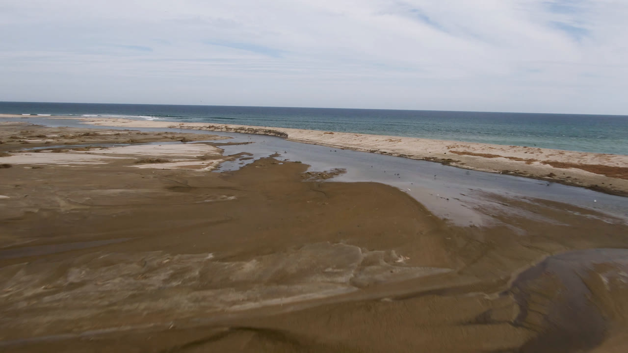 Aerial frame in San Jose del Cabo with wet sand flats in the foreground, a narrow stream through the center, pale beach dunes beyond, and ocean surf under scattered clouds.