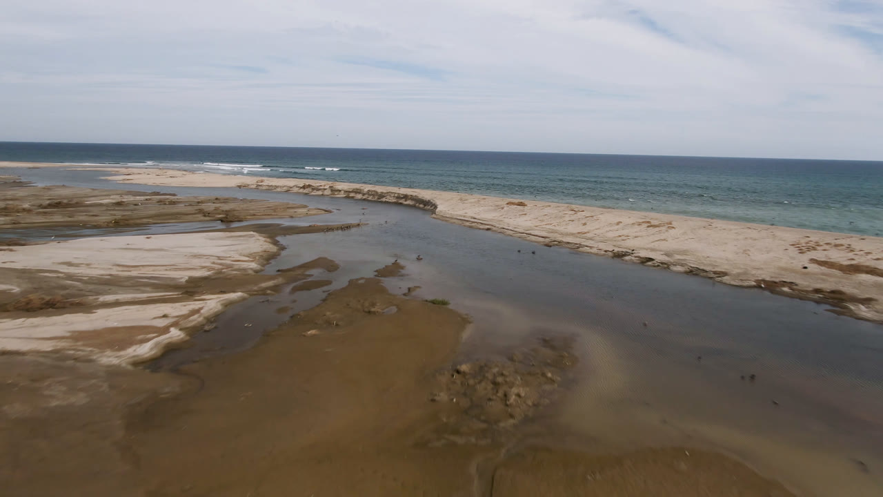 Aerial view in San Jose del Cabo showing a shallow stream and dark wet sand in the foreground, pale sand banks in the midground, ocean surf beyond, and no visible people.