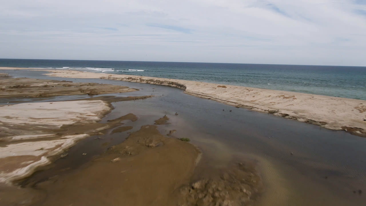 Aerial frame from San Jose del Cabo with the stream channel centered, a pale sand spit curving across the beach, blue-green ocean in the distance, and no visible people.