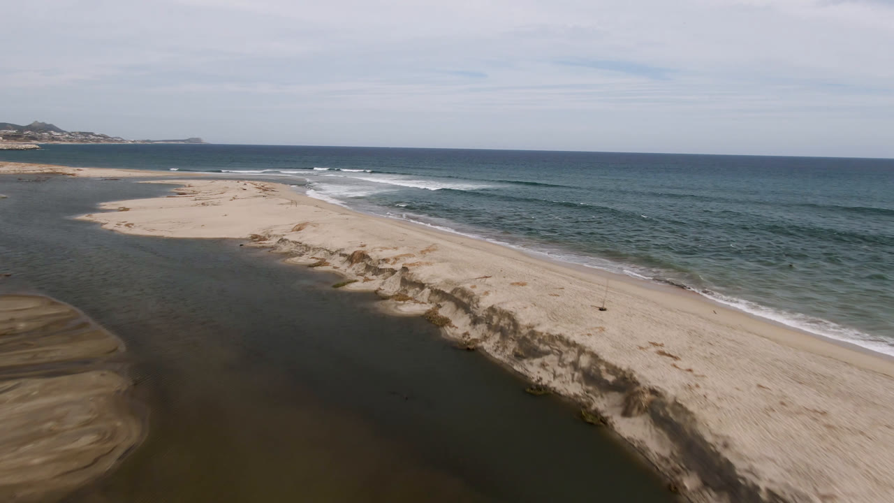 Aerial frame over San Jose del Cabo showing a pale sand spit between dark lagoon water and blue ocean surf, with distant coast and no visible people.