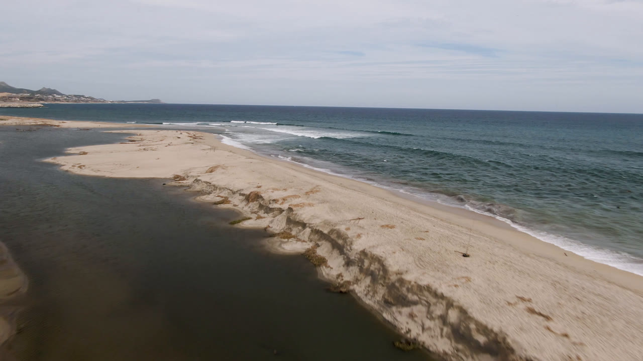 Drone frame over San Jose del Cabo with the lagoon widening at left, pale beach through the center, blue ocean at right, and no visible people on the sand.