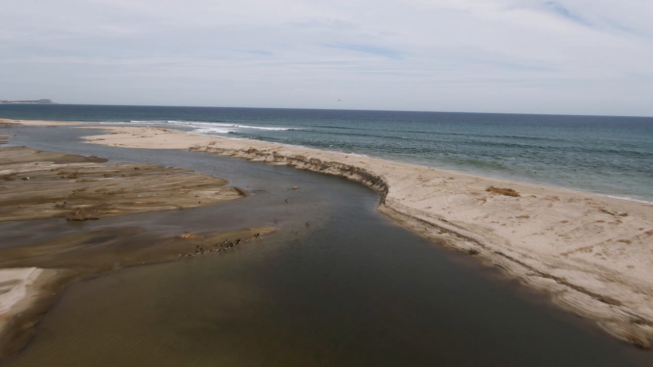 Aerial frame over San Jose del Cabo sand banks, with a dark stream bending toward the ocean, exposed wet flats on the left, surf on the right, and no visible people.