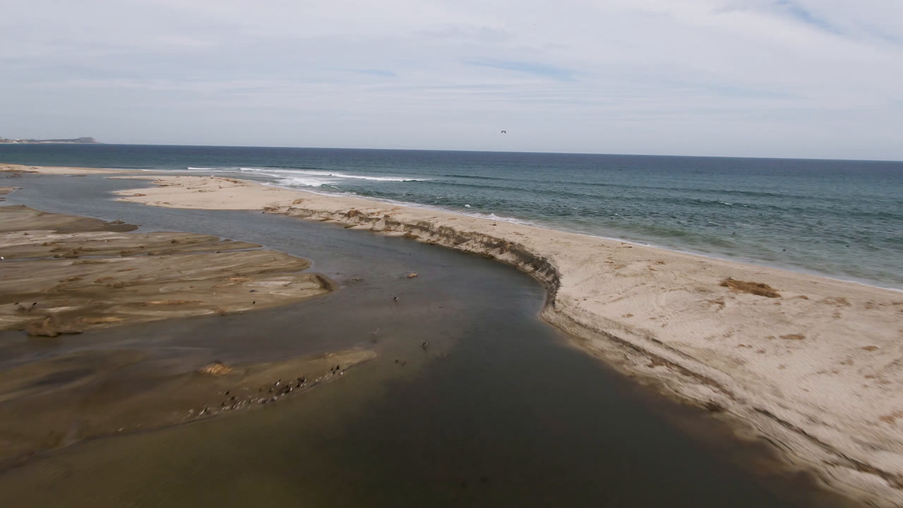Aerial view in San Jose del Cabo showing the stream channel wider in the foreground, pale sand ridge along the shore, blue ocean beyond, and no visible people.