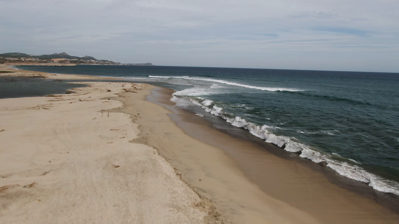 Wide San Jose del Cabo shoreline frame with a pale sand spit in the center, breaking surf along the right edge, distant coast on the horizon, and no visible people.