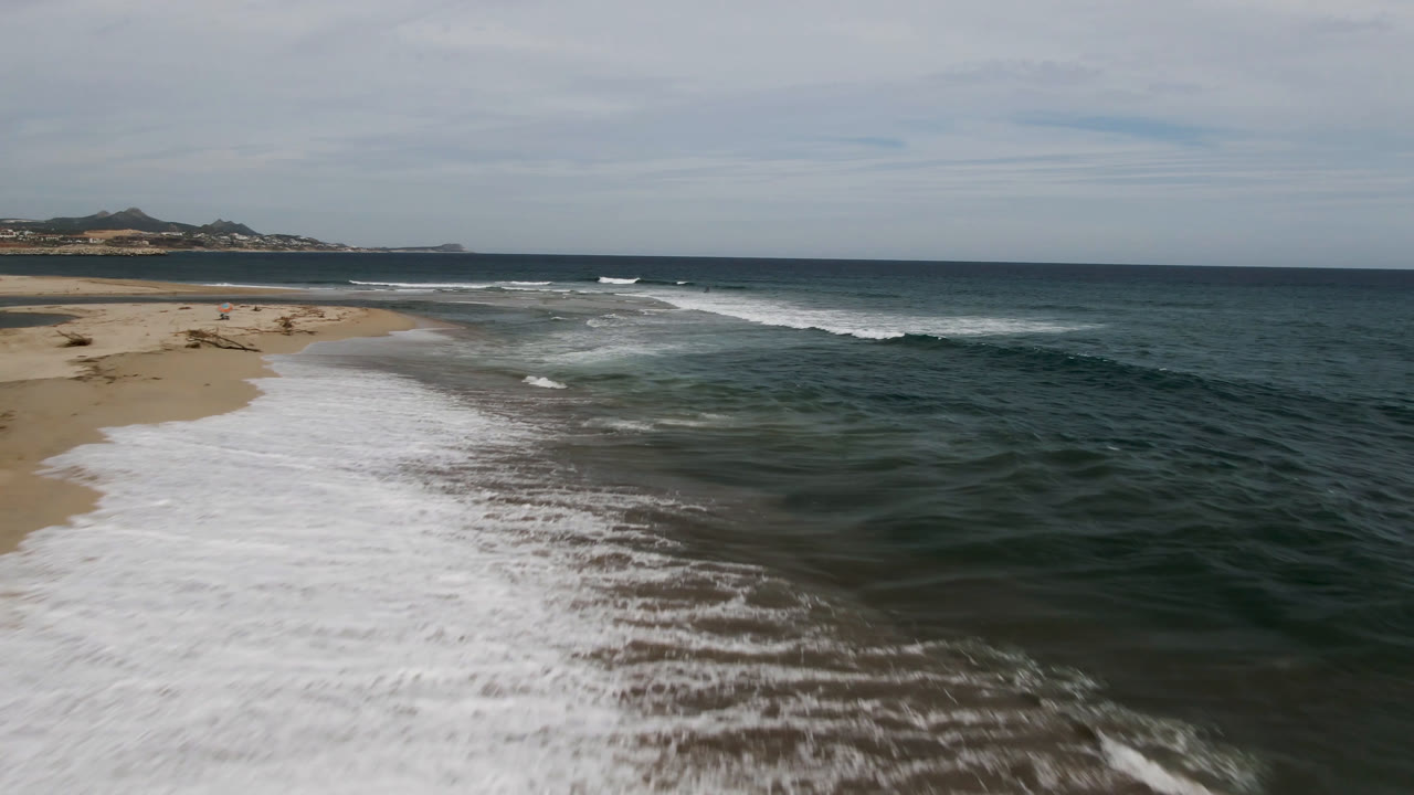 Aerial view of surf spreading across a sandy beach in San Jose del Cabo, with dark green ocean to the right, distant land on the horizon, and no visible people.