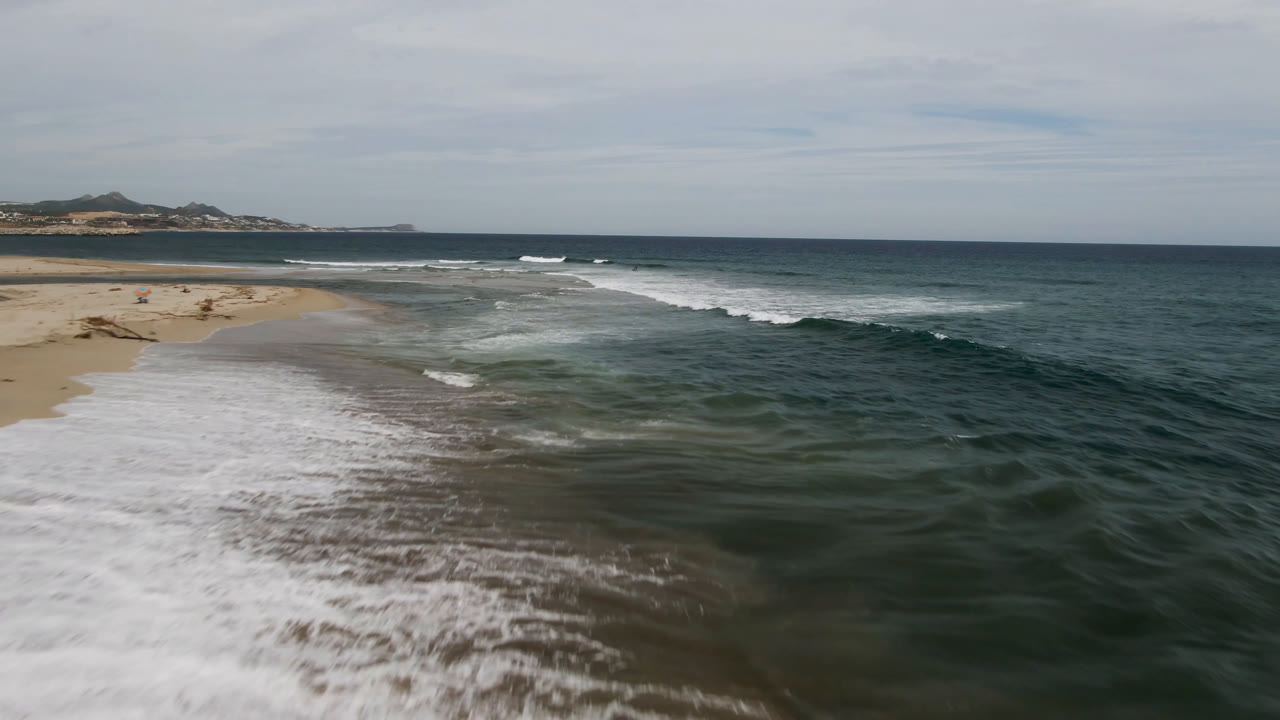 Aerial frame of a curving sand edge in San Jose del Cabo as a broad sheet of white foam moves inland, with offshore breakers and no visible people.