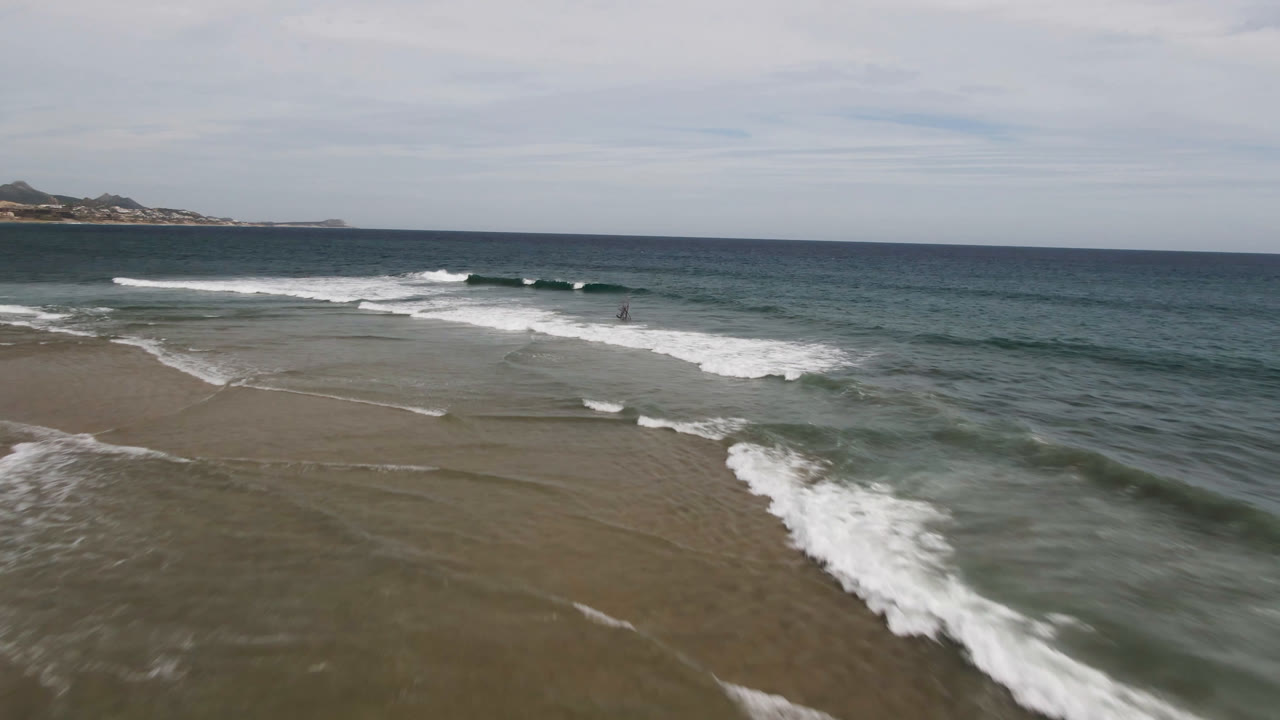 Aerial frame from San Jose del Cabo showing white surf curling over shallow wet sand, green-blue ocean beyond, distant land on the horizon, and no visible people.