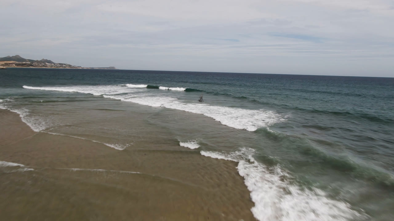 Aerial San Jose del Cabo beach view with a broad white wave band crossing the shallow water, darker ocean behind it, distant land near the horizon, and no visible people.