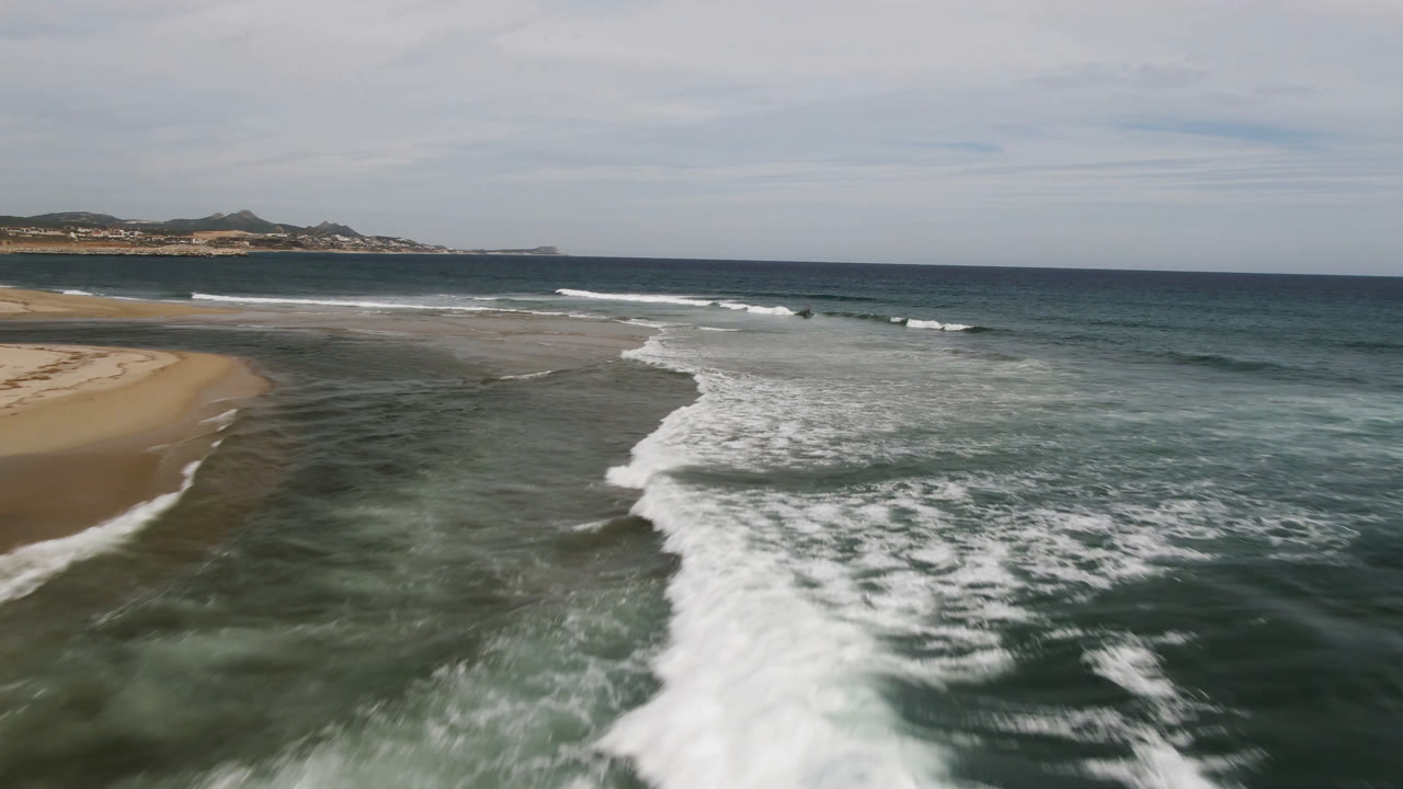 Aerial view over San Jose del Cabo surf, with foamy water in the foreground, sandy beach on the left, distant land on the horizon, and no visible people.