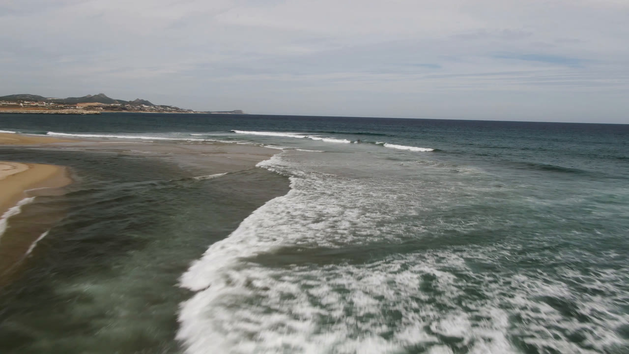 Aerial frame from San Jose del Cabo showing a curved white surf line spreading toward the lower frame, sand at left, blue-green ocean at right, and no visible people.
