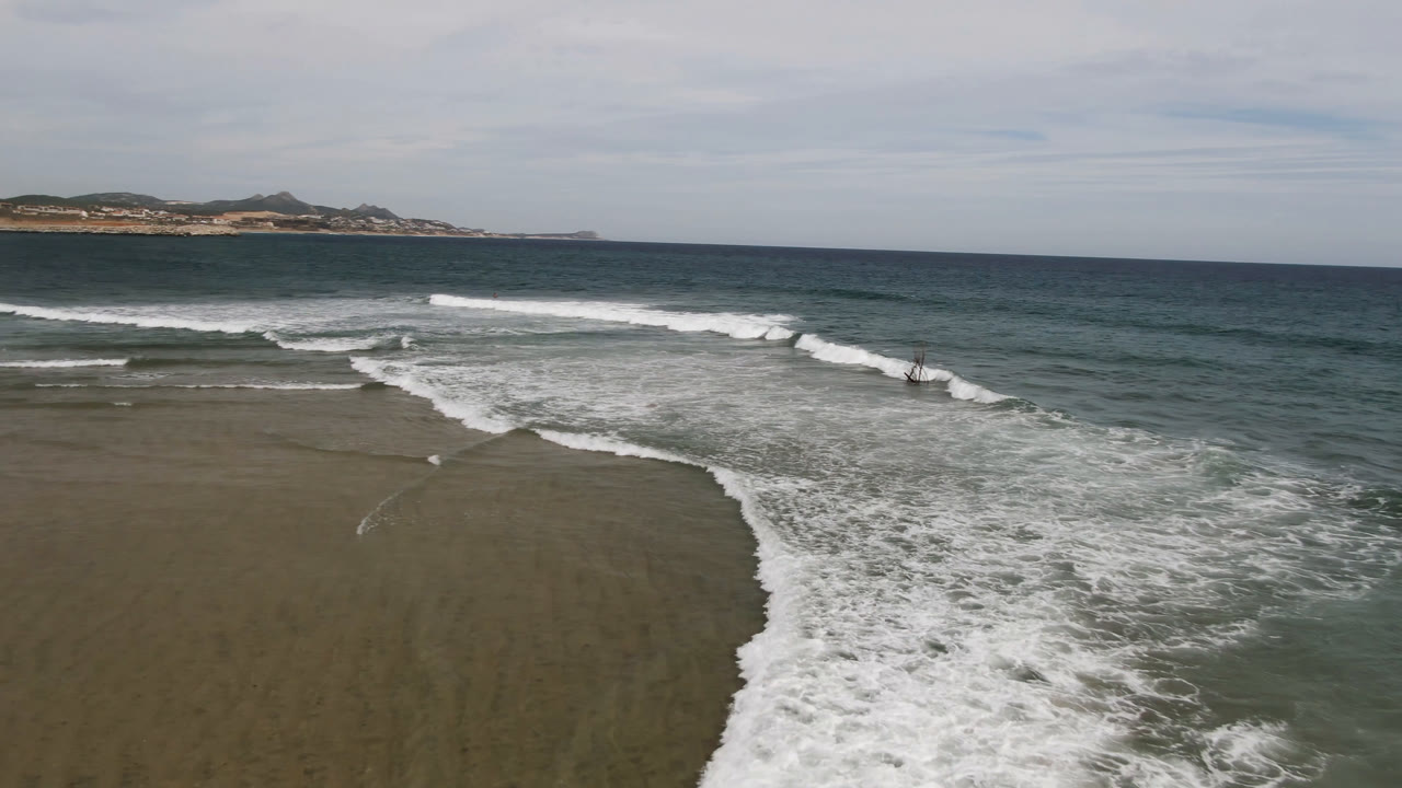 Aerial frame over San Jose del Cabo shoreline with white surf curling across wet sand, green-blue ocean beyond, distant low hills, clouded sky, and no visible people.