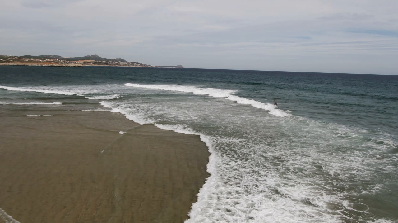 Aerial view in San Jose del Cabo with a broad foamy wave band sliding across the beach, darker ocean water in the background, distant shoreline, and no visible people.