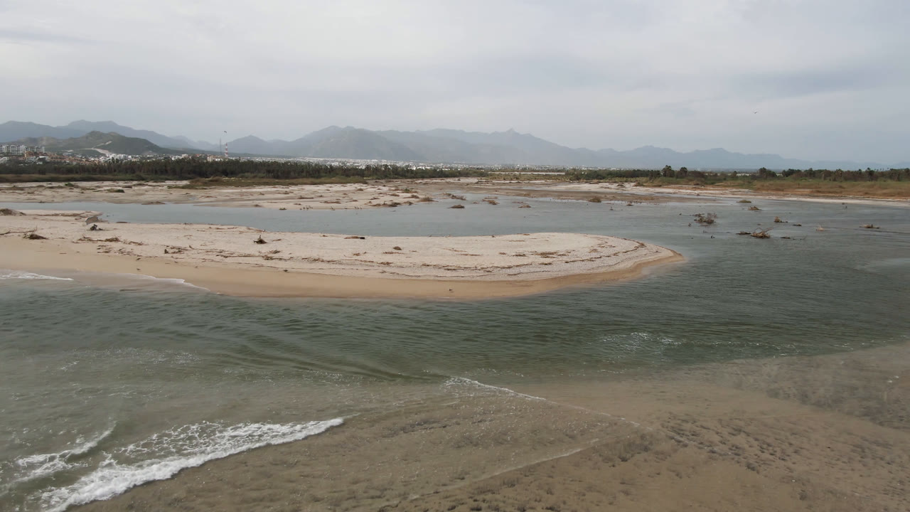 At 0:00, an aerial view in San Jose del Cabo shows shallow surf in the foreground, a pale sandbar and lagoon in the middle distance, hazy mountains beyond, and no visible people.